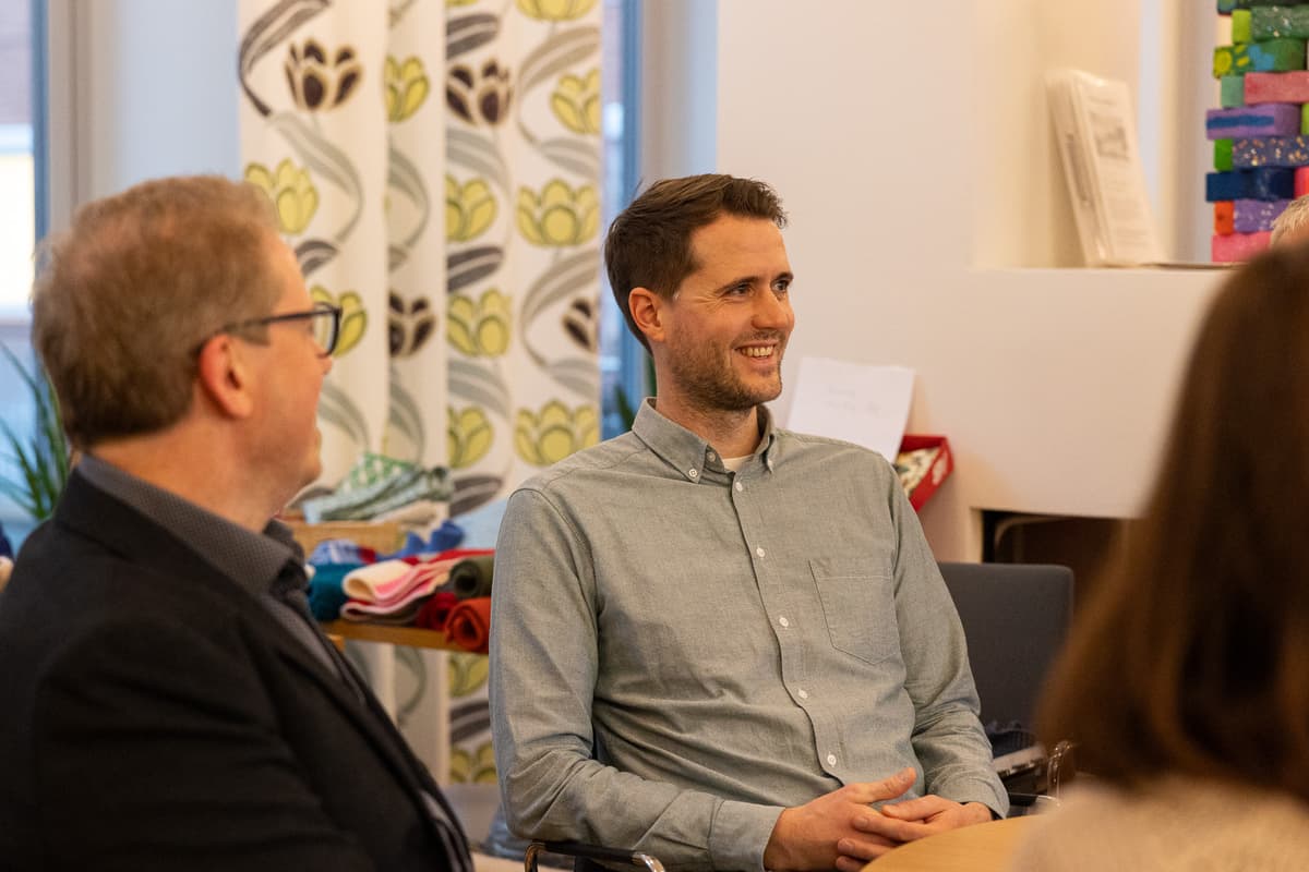 Yngve Skålnes En blidspent mann sitter ved et bord. Vi ser profilen til en annen smilende mann.