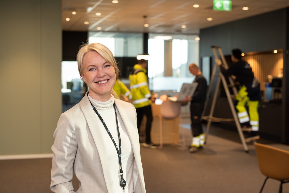 Camilla Berge Jacobsen 2 Smilende kvinne foran folk i arbeid på en gardintrapp og rundt et bord.