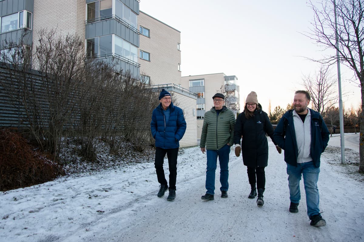 Energikartlegging elvebredden Tre menn og en dame, alle vinterkledde, går på fortauet utenfor et borettslag med mursteinsblokker.